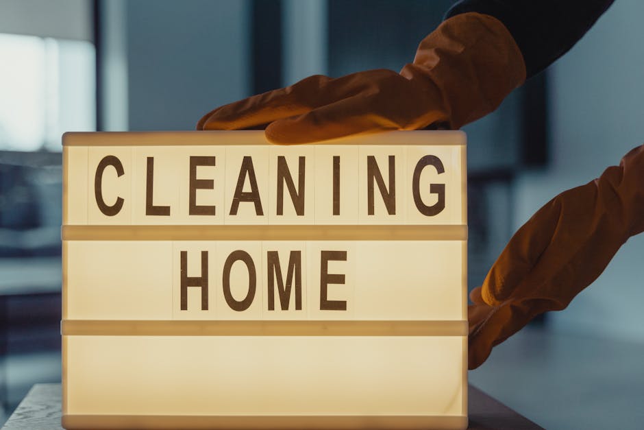Close-up of a person in pink rubber gloves using a spray bottle of cleaning solution to disinfect a white windowsill in a kitchen. The windowsill has a small potted green plant, and a blue cleaning cl